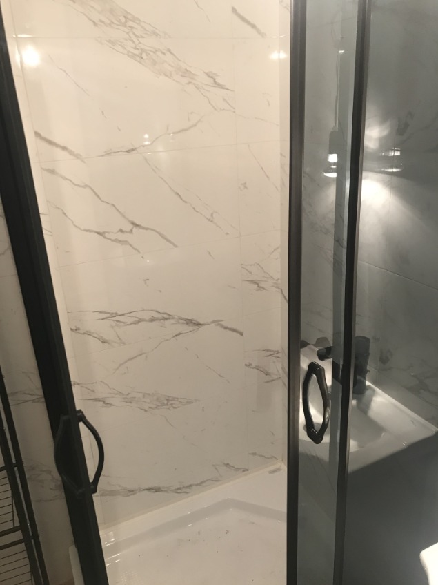 Kabina prysznicowa z czarnymi profilami i uchwytami, białe płytki imitujące marmur na ścianach, biały brodzik.