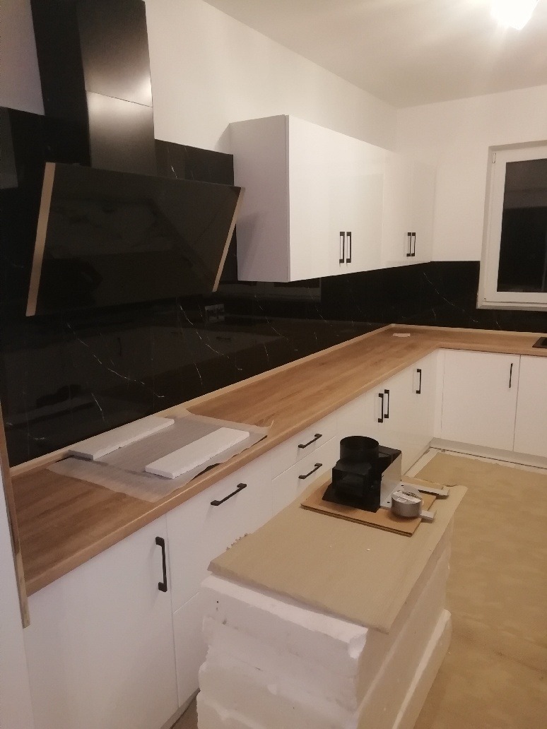 Nowoczesna kuchnia w trakcie montażu: białe szafki z czarnymi uchwytami, drewniany blat, czarny okap z marmurkowym wzorem na ścianie, widoczne elementy montażowe na blacie.