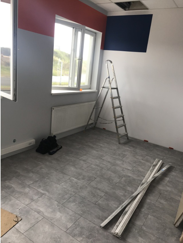 Przygotowane pomieszczenie biurowe do malowania: ściany częściowo pomalowane na szaro, czerwono i granatowo, drabina aluminiowa, listwy przypodłogowe i torba narzędziowa na podłodze z szarych płytek.