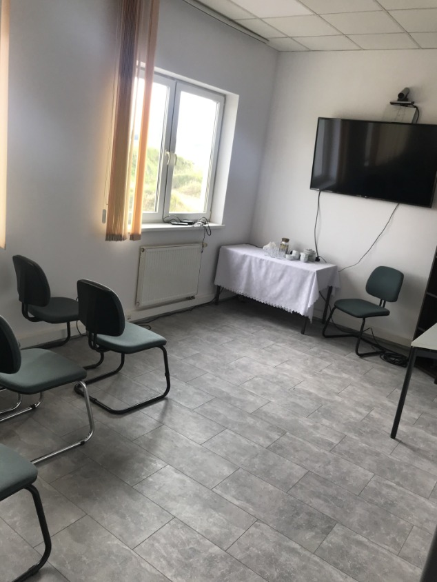 Szare płytki podłogowe imitujące drewno w jasnym biurze z krzesłami, stołem nakrytym białym obrusem i telewizorem na ścianie.