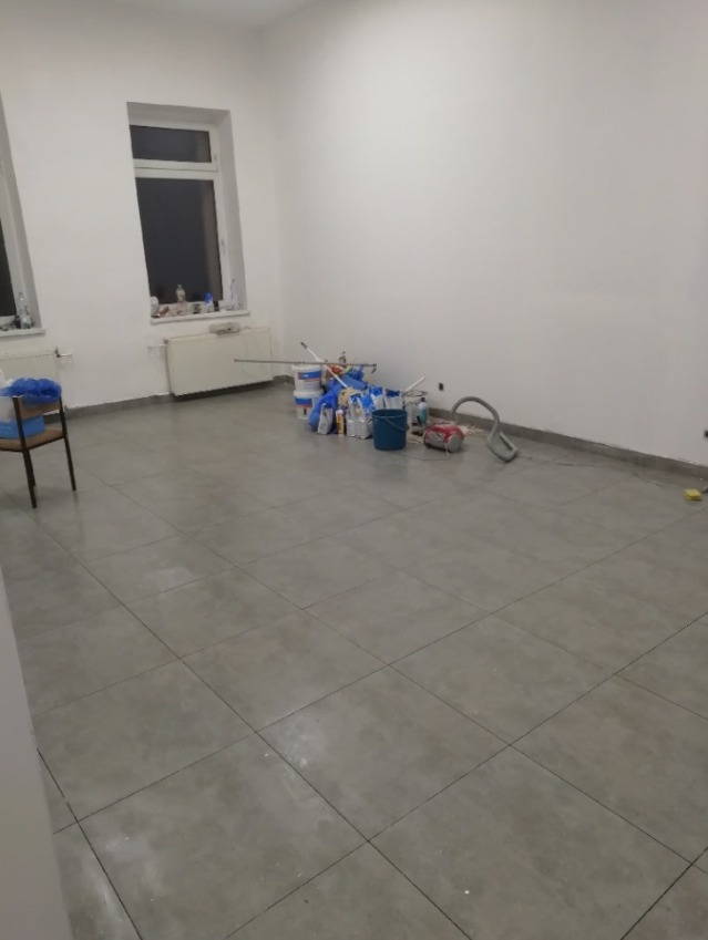 Pokój z szarymi płytkami podłogowymi, białe ściany, widoczne pędzle malarskie, wałki, wiadra z farbą i inne akcesoria malarskie przygotowane do pracy.