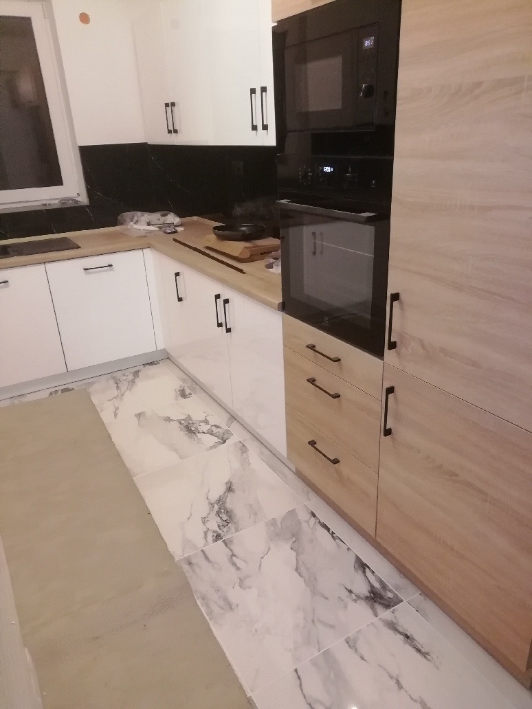 Nowoczesna kuchnia z białymi i drewnopodobnymi szafkami, czarnym panelem nad blatem i marmurową podłogą, z zabudowanym piekarnikiem i mikrofalówką.