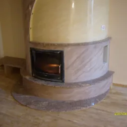 Okrągły kominek z marmurową obudową, z palącym się wewnątrz ogniem, w pokoju z drewnianą podłogą i kremowymi ścianami.