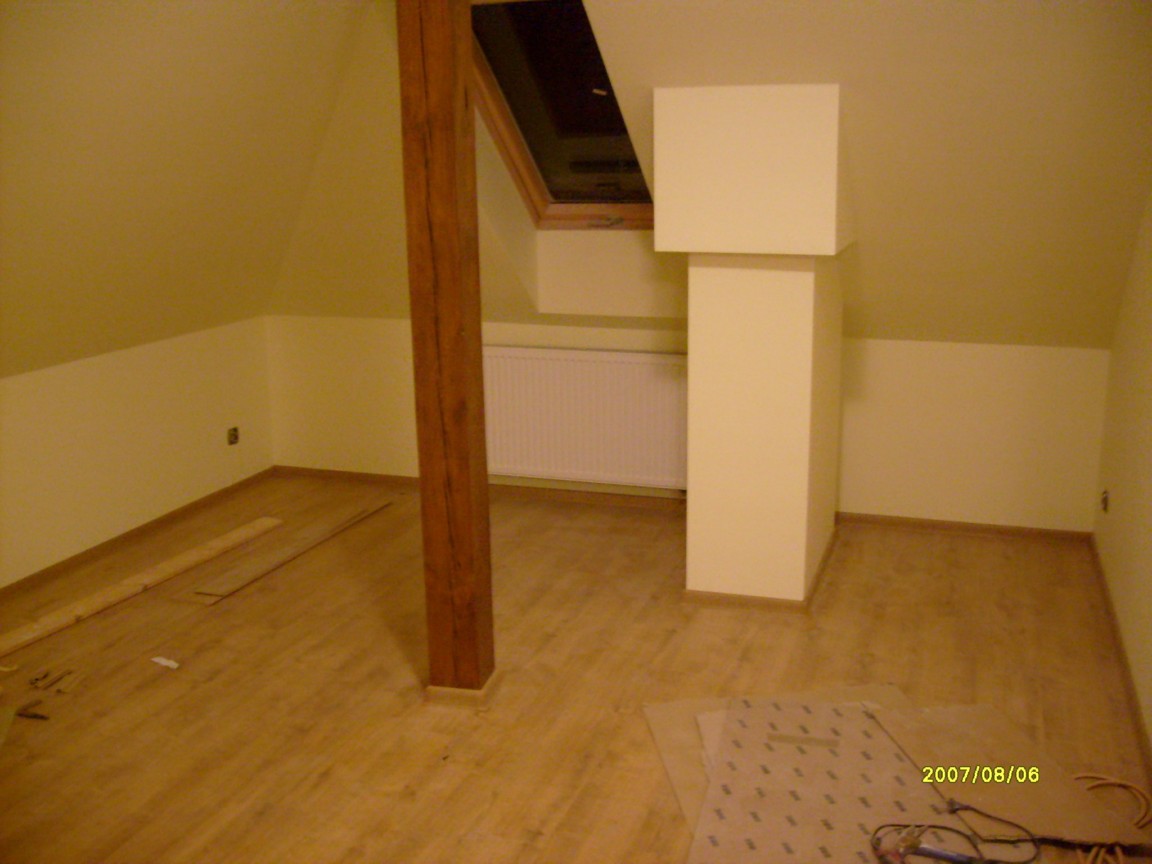 Poddasze po remoncie z drewnianą podłogą, beżowymi ścianami, drewnianą belką konstrukcyjną i obudowaną rurą wentylacyjną. Widoczne narzędzia i materiały budowlane.