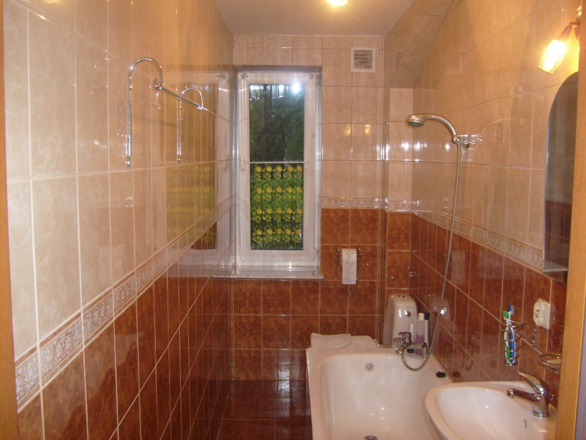 Jasna łazienka z beżowo-brązowymi płytkami, białą wanną i umywalką, widok na zieleń za oknem, chromowana armatura i akcesoria.