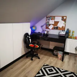 Pokój na poddaszu ze skośnym sufitem, białą szafą w zabudowie, czarnym biurkiem, krzesłem gamingowym z arbuzem zamiast siedziska, korkową tablicą i dywanem w geometryczne wzory.