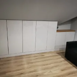 Biała, lakierowana zabudowa meblowa pod skosem na poddaszu, z widocznym fragmentem czarnej sofy i jasną podłogą imitującą drewno.