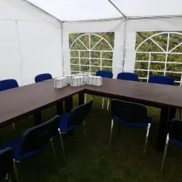 Wnętrze białego namiotu z ciemnobrązowym stołem w kształcie litery L, niebieskimi krzesłami i dekoracyjnymi szklankami na stole, widok na zieleń przez okna.