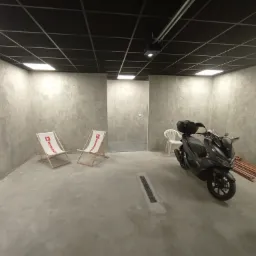 Nowoczesne wnętrze garażu z szarymi ścianami o surowym wykończeniu, czarnym sufitem kasetonowym z oświetleniem LED, dwoma leżakami z logo Rockwool, motocyklem i ułożonymi deskami przy ścianie.