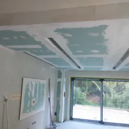 Sufit z płyt kartonowo-gipsowych w trakcie montażu, widoczne szpachlowane łączenia i wnęki na oświetlenie, ściana z zamontowaną płytą, okno balkonowe z widokiem na zieleń.