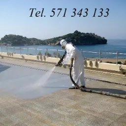 Pracownik w kombinezonie ochronnym natryskuje szarą substancję na płaski dach z widokiem na morze i wyspę w oddali, widoczny numer telefonu na zdjęciu.