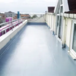 Wykończony płaski dach budynku wielorodzinnego z szarą, gładką powierzchnią hydroizolacyjną i metalową balustradą, widok na miasto i morze w tle.