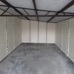 Wnętrze szopy z metalową konstrukcją i ścianami pokrytymi pianką izolacyjną, widoczne ślady montażu i szara posadzka.