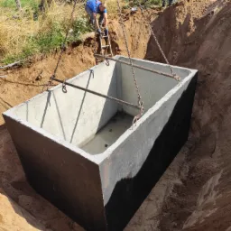 Betonowy zbiornik, prawdopodobnie szambo, opuszczany do wykopu za pomocą dźwigu, widoczni pracownicy nadzorujący proces