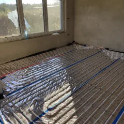 Widok na rozłożoną instalację ogrzewania podłogowego na folii izolacyjnej w surowym pomieszczeniu z oknem, widoczne białe rurki grzewcze i niebieskie taśmy mocujące.