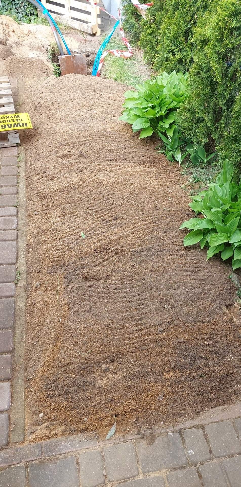 Świeżo zasypany piaskiem wykop w ogrodzie, zabezpieczony taśmą ostrzegawczą, z widocznymi narzędziami i tabliczką informującą o głębokim wykopie, obok rosną rośliny.