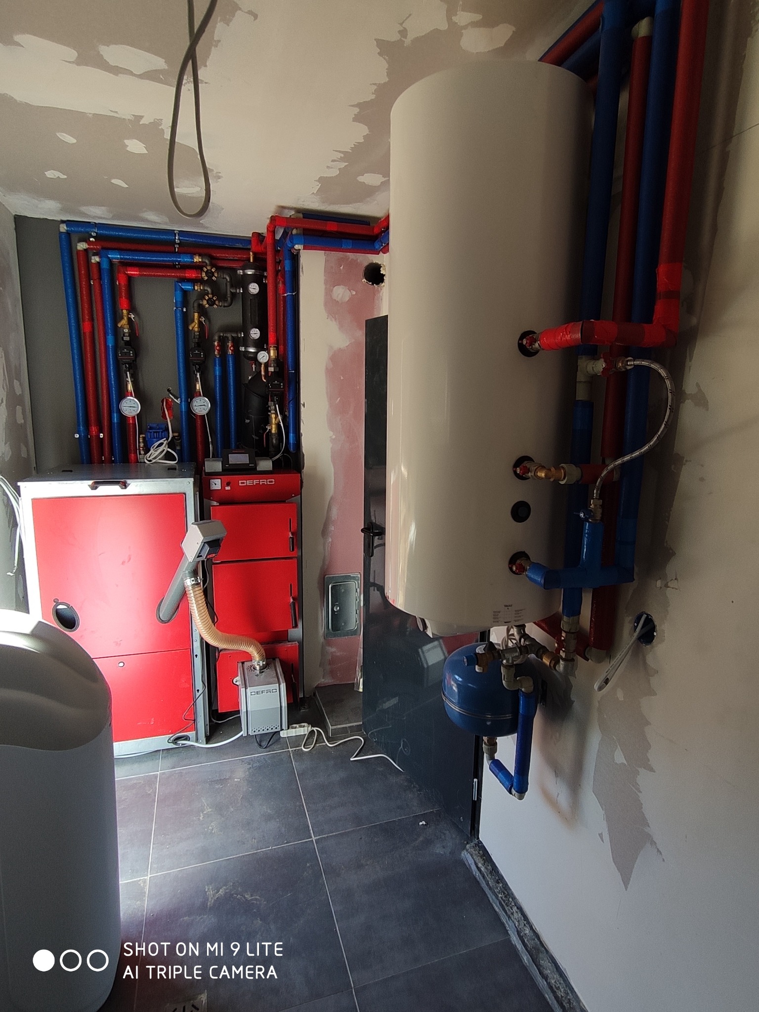 Wnętrze kotłowni w trakcie montażu: czerwony piec Defro z podajnikiem, biały zasobnik ciepłej wody i instalacja z rur w kolorach czerwonym i niebieskim.