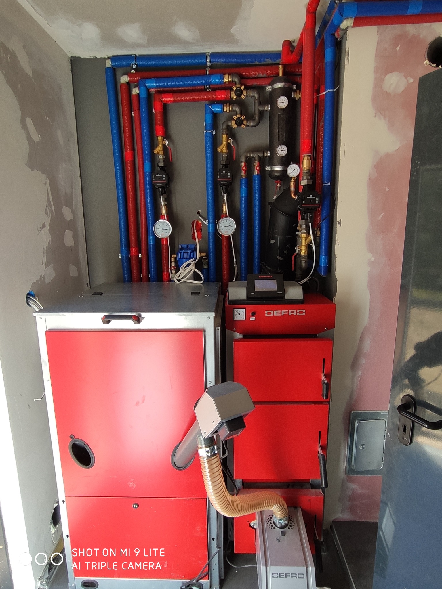 Czerwony piec CO marki Defro z podajnikiem ślimakowym i rurami instalacji grzewczej w kolorach czerwonym i niebieskim, zamontowany w pomieszczeniu w trakcie wykańczania.