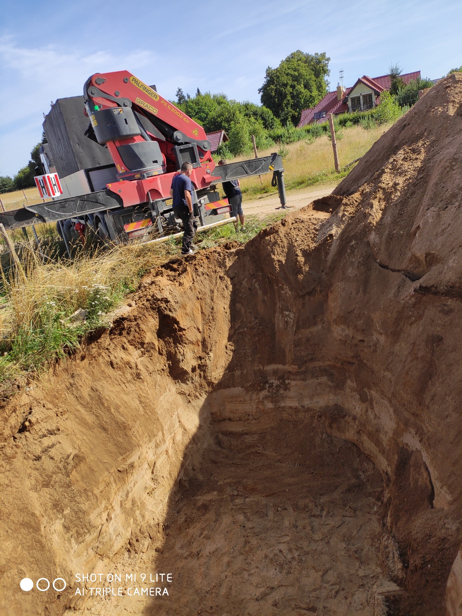 Głęboki wykop w ziemi przygotowany pod instalację, z widocznym dźwigiem hydraulicznym Palfinger i dwoma pracownikami nadzorującymi prace. W tle dom z czerwoną dachówką i zielona roślinność.