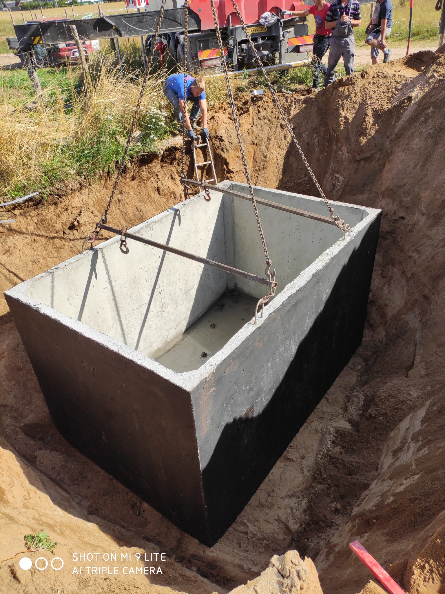 Betonowy zbiornik, prawdopodobnie szambo, opuszczany do wykopu za pomocą dźwigu, widoczni pracownicy nadzorujący proces