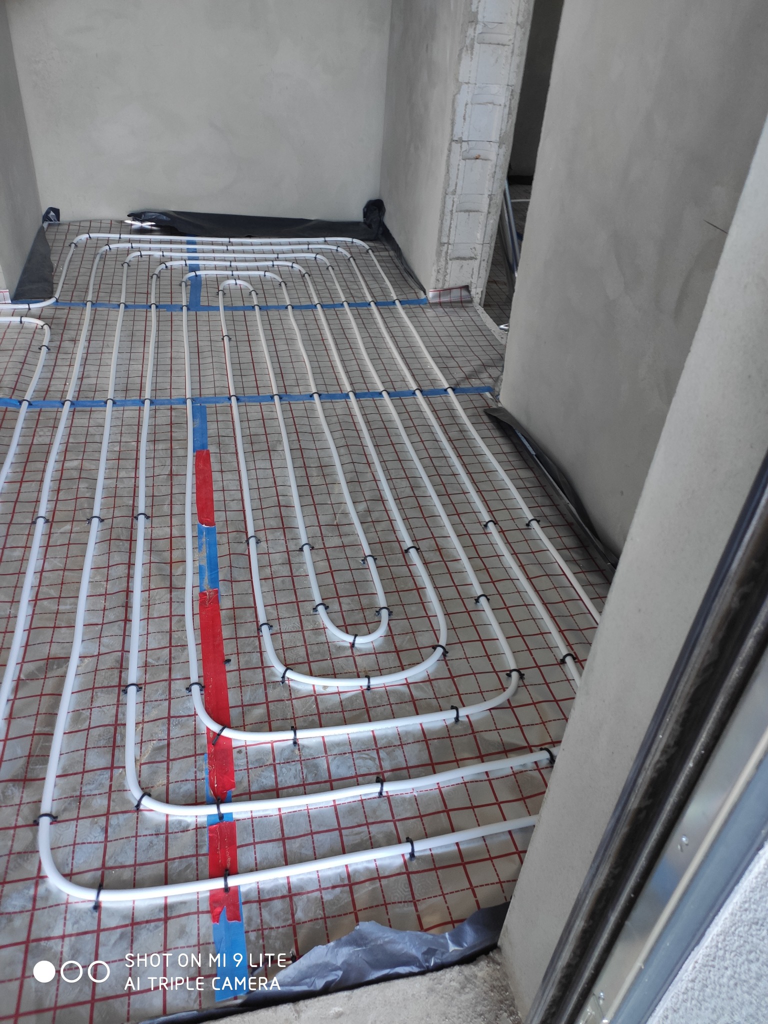 Białe rurki ogrzewania podłogowego, zamocowane czarnymi klipsami na siatce z czerwoną kratą, częściowo przykryte czarną folią, w surowym wnętrzu przed wylewką.