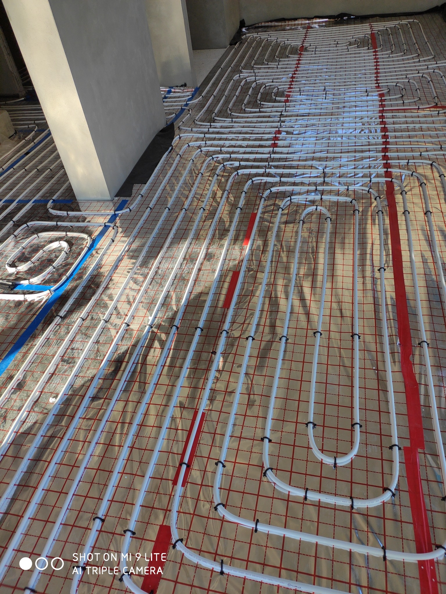 Białe rury ogrzewania podłogowego ułożone na folii z czerwoną kratką, mocowane czarnymi klipsami, widoczne fragmenty niebieskiej taśmy i betonowy filar.