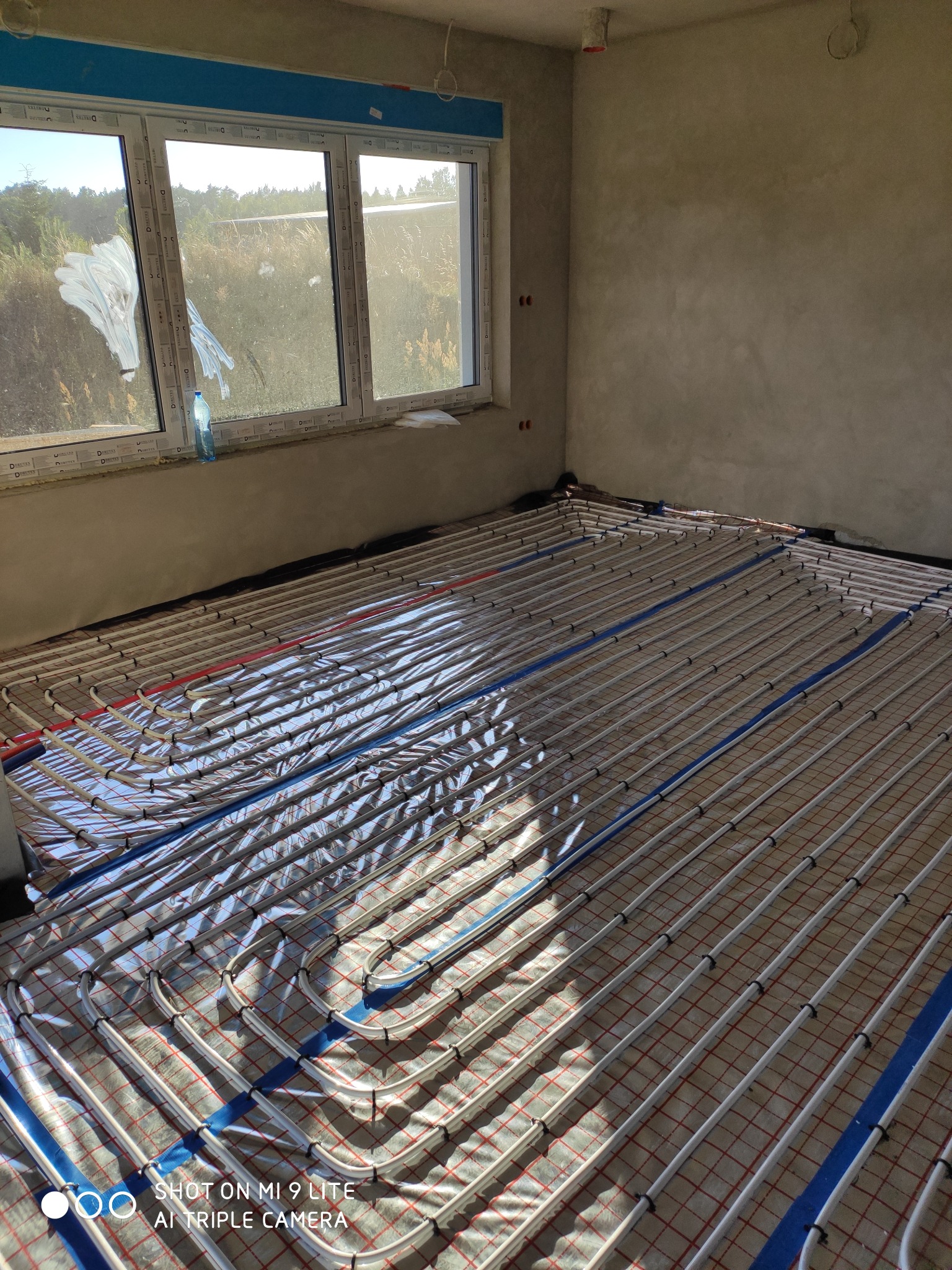 Widok na rozłożoną instalację ogrzewania podłogowego na folii izolacyjnej w surowym pomieszczeniu z oknem, widoczne białe rurki grzewcze i niebieskie taśmy mocujące.