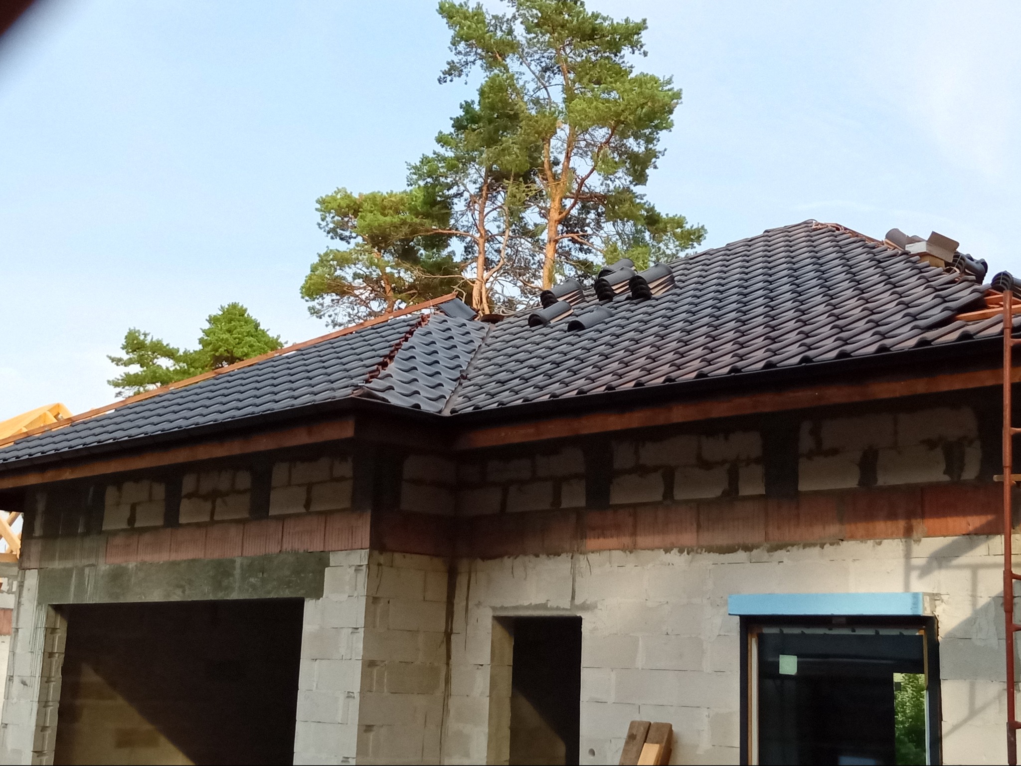 Budowa domu jednorodzinnego w trakcie. Widoczny fragment dachu pokrytego ciemną dachówką ceramiczną oraz surowy stan ścian z betonu komórkowego. Na dachu leżą jeszcze niezamontowane dachówki, a...
