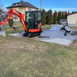Pomarańczowa mini koparka Kubota z zamontowanym specjalistycznym chwytakiem na tle ułożonej kostki brukowej, przyczepy i domu jednorodzinnego.