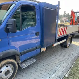 Niebieski samochód dostawczy z przyczepą przewożącą małą koparkę, zaparkowany na brukowanej ścieżce.