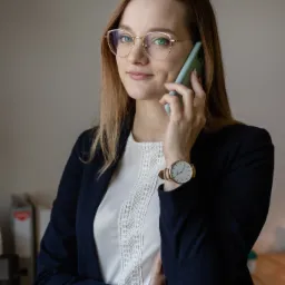 Profesjonalnie ubrana kobieta w okularach, rozmawia przez telefon, nosząc złoty zegarek na nadgarstku.