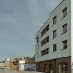 Nowoczesny budynek biurowy z ciemną cegłą na parterze i jasną elewacją, widoczny z ulicy w pochmurny dzień, z reklamą na przystanku autobusowym w tle.