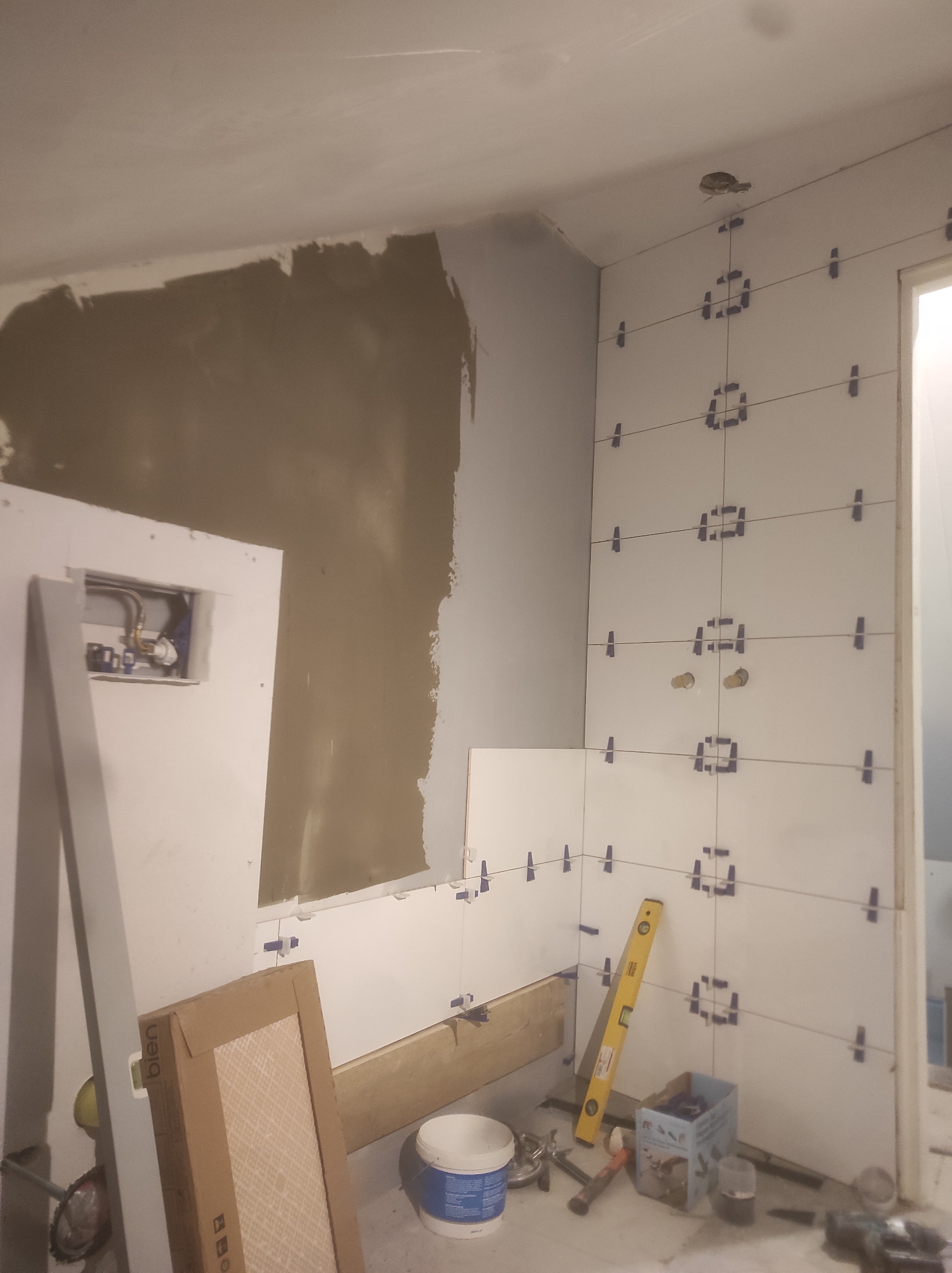 Ściana łazienki w trakcie układania białych płytek, z użyciem krzyżyków dystansowych i poziomicy, widoczne przygotowane podłączenia hydrauliczne, fragment ściany z nałożonym gruntem.