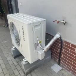 Zewnętrzna jednostka pompy ciepła Kaisai zamontowana na betonowych podstawach przy szarej ścianie budynku z widocznymi przyłączami hydraulicznymi i odprowadzeniem skroplin w srebrnej izolacji.