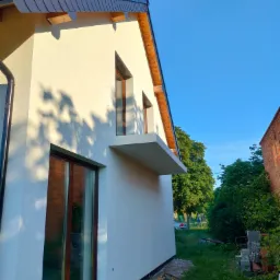 Boczny widok otynkowanej, jasnej elewacji domu jednorodzinnego z brązowymi oknami, widoczne cienie drzew na ścianie i fragment dachu z czarną dachówką