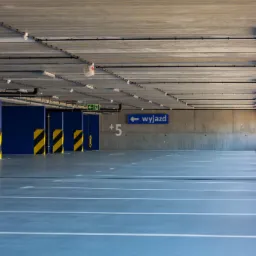 Wnętrze parkingu wielopoziomowego z szarą posadzką, białymi liniami wyznaczającymi miejsca parkingowe, żółto-czarnymi pasami na kolumnach i betonowym sufitem z instalacjami.