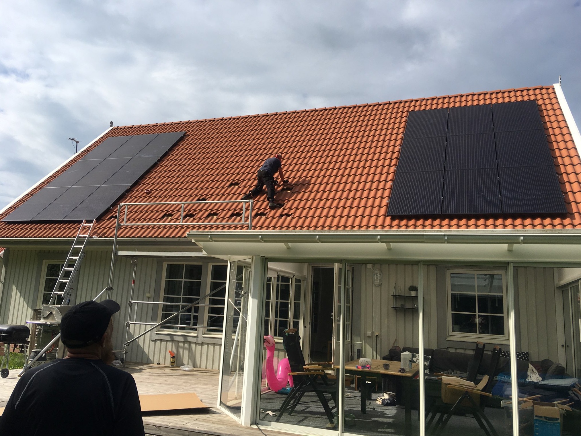 Montaż paneli słonecznych na dachu domu z czerwonej dachówki; widoczny instalator na dachu, drabina i częściowo widoczny mężczyzna obserwujący prace z dołu.