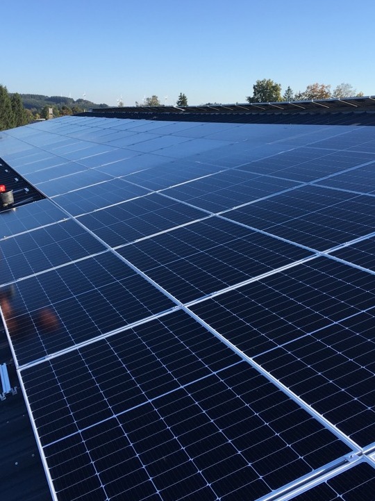 Rząd paneli słonecznych z widocznym odbiciem nieba, zainstalowanych na dachu domu z widocznymi elementami montażowymi i narzędziem w tle, w słoneczny dzień.