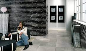 Kobieta siedzi na pufie w nowoczesnym wnętrzu z czarną ceglaną ścianą, jasną podłogą i minimalistycznymi dekoracjami ściennymi.