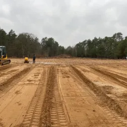Żółta koparka gąsienicowa i zagęszczarka wibracyjna na wyrównanym, piaszczystym terenie budowy, widoczne ślady opon, w tle las iglasty pod pochmurnym niebem.