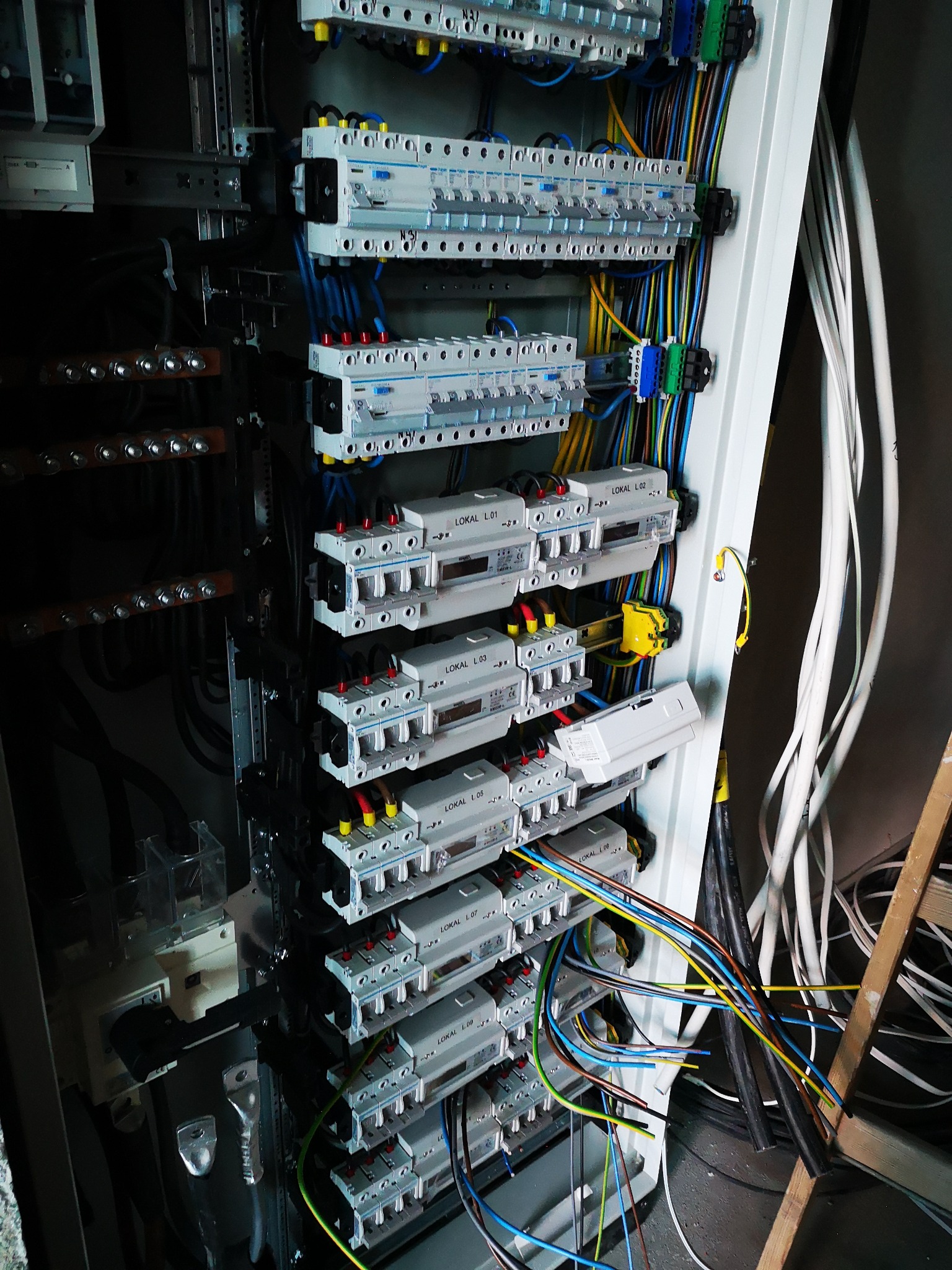 Rozdzielnica elektryczna z zainstalowanymi bezpiecznikami i okablowaniem, widoczne oznaczenia LOKAL L.01, L.02, L.03, L.07, L.08, L.09, kable w kolorach niebieskim, żółtym, zielonym, brązowym...