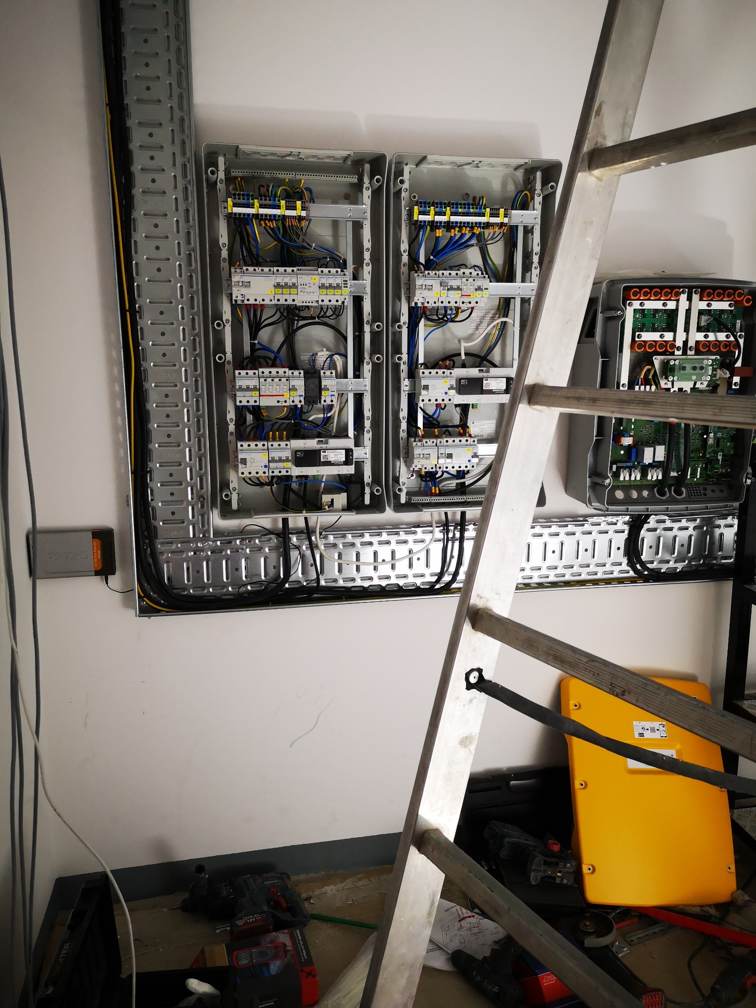 Dwa otwarte panele rozdzielcze na ścianie, z widocznymi przewodami i elementami elektrycznymi, obok drabina aluminiowa i narzędzia na podłodze.