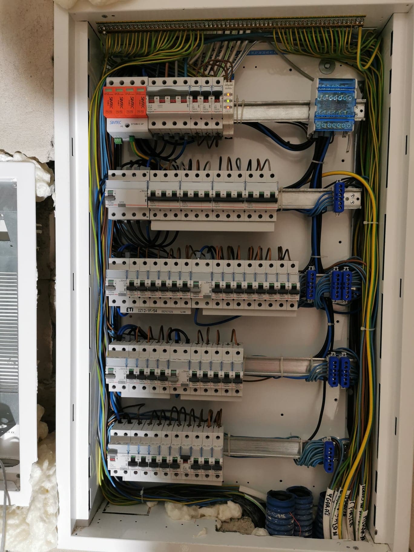 Rozdzielnica elektryczna z licznymi zabezpieczeniami, przewodami oznaczonymi opisami, zamontowana w białej obudowie, widoczne zabezpieczenia przeciwprzepięciowe SIMTEC i wyłączniki nadprądowe Legrand.