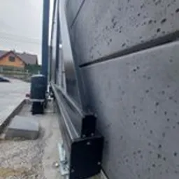Szara, przesuwna brama wjazdowa z widocznym mechanizmem automatycznego otwierania i betonowym fundamentem, zamontowana przy nowoczesnym ogrodzeniu z betonu architektonicznego.