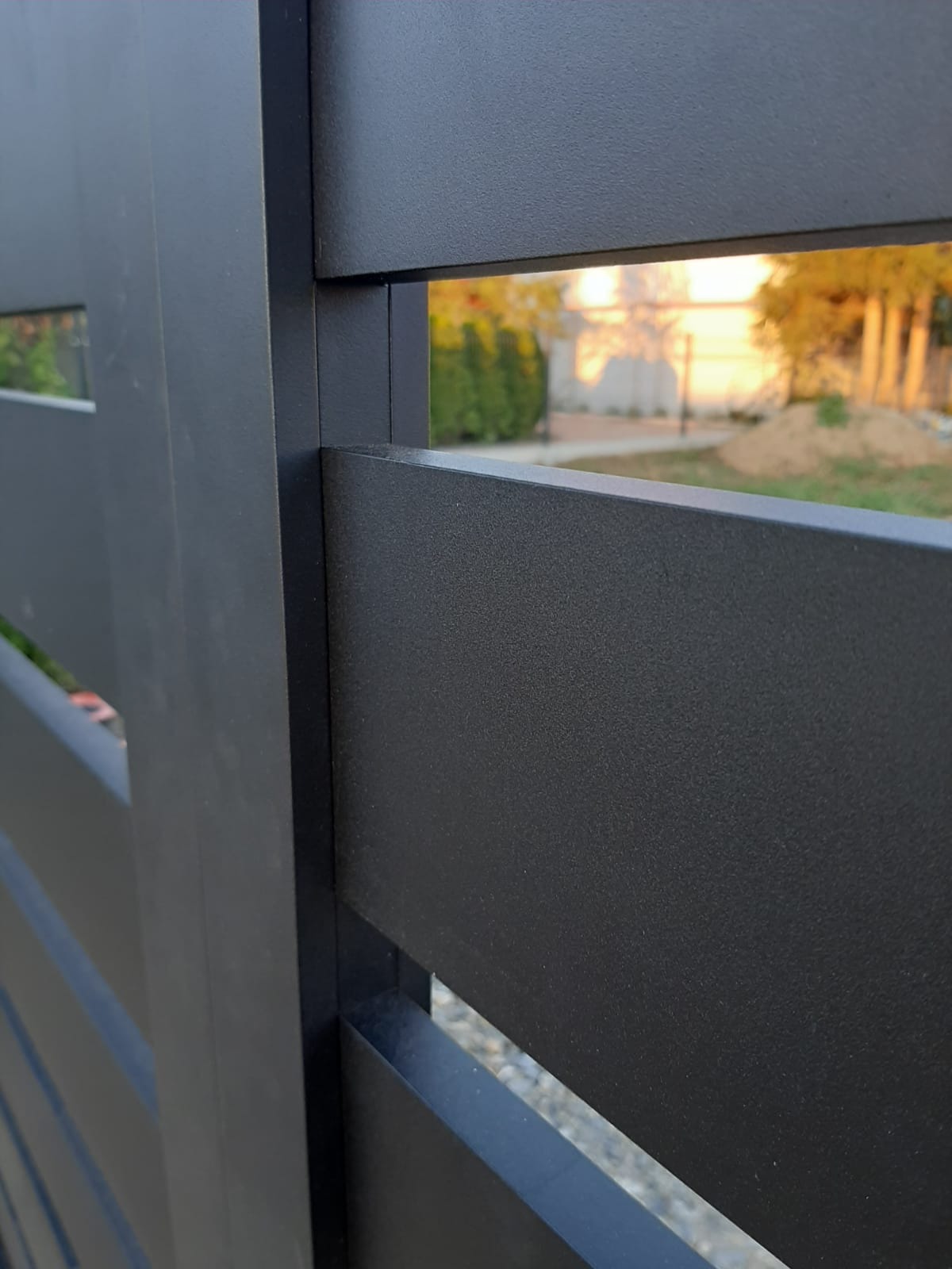 Fragment nowoczesnego, matowego, grafitowego ogrodzenia metalowego z poziomymi panelami i wąskimi prześwitami, z widokiem na ogród w tle.