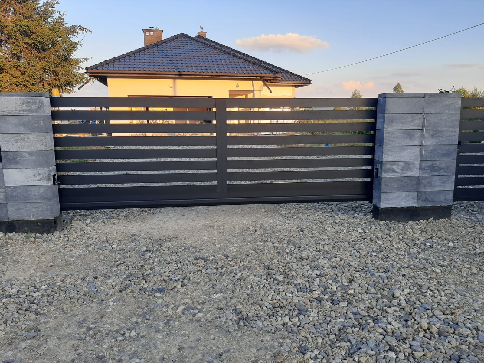 Automatyczna brama przesuwna z poziomymi panelami, zamontowana na posesji z domem w tle, widoczna z poziomu gruntu pokrytego żwirem.