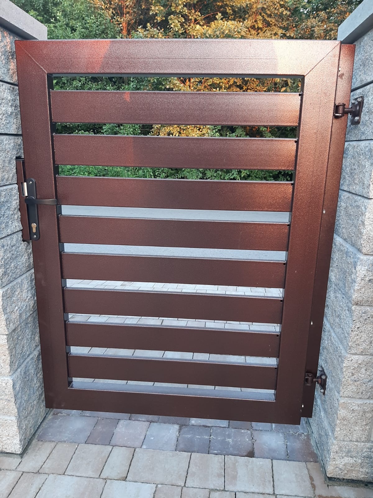 Nowoczesna, brązowa furtka z poziomych profili, zamontowana na kamiennych słupkach, widok z bliska na zamek i zawiasy, kostka brukowa na podłożu, zieleń w tle.