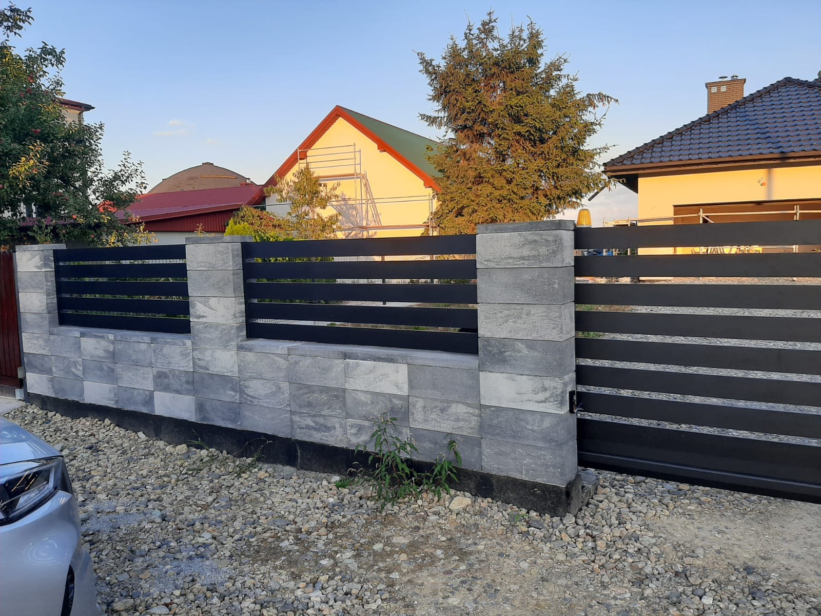 Nowoczesne ogrodzenie z szarej kostki i czarnych, poziomych przęseł, brama przesuwna w tle, widok z poziomu gruntu na tle domów.