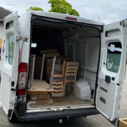 Otwarty tył białego furgonetki załadowany stołami i krzesłami owiniętymi folią stretch, na zewnątrz widoczne drewniane panele ogrodzeniowe i kostka brukowa.