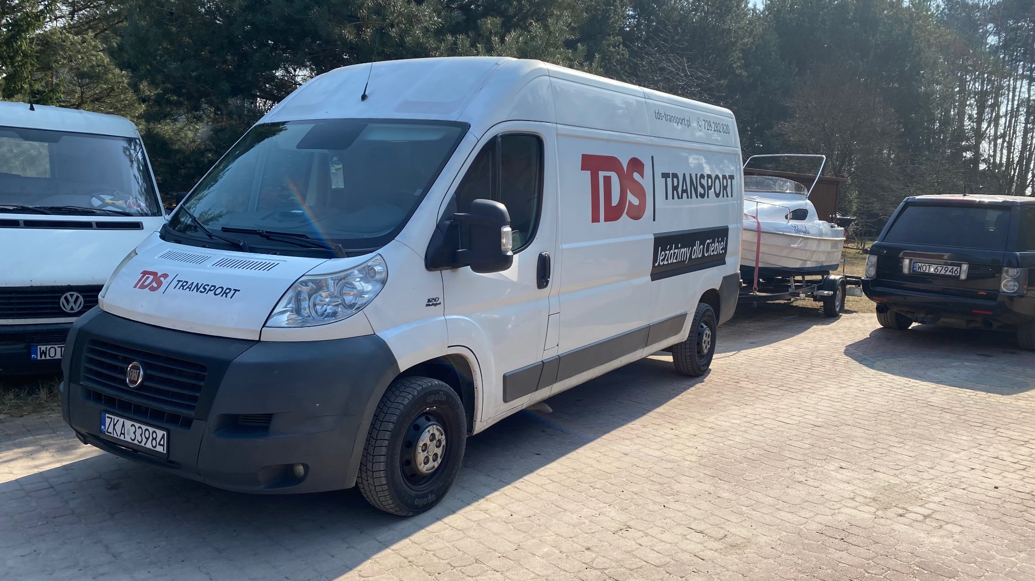 Biały samochód dostawczy z logo firmy transportowej TDS ciągnie przyczepę z łodzią na tle drzew i drugiego vana.